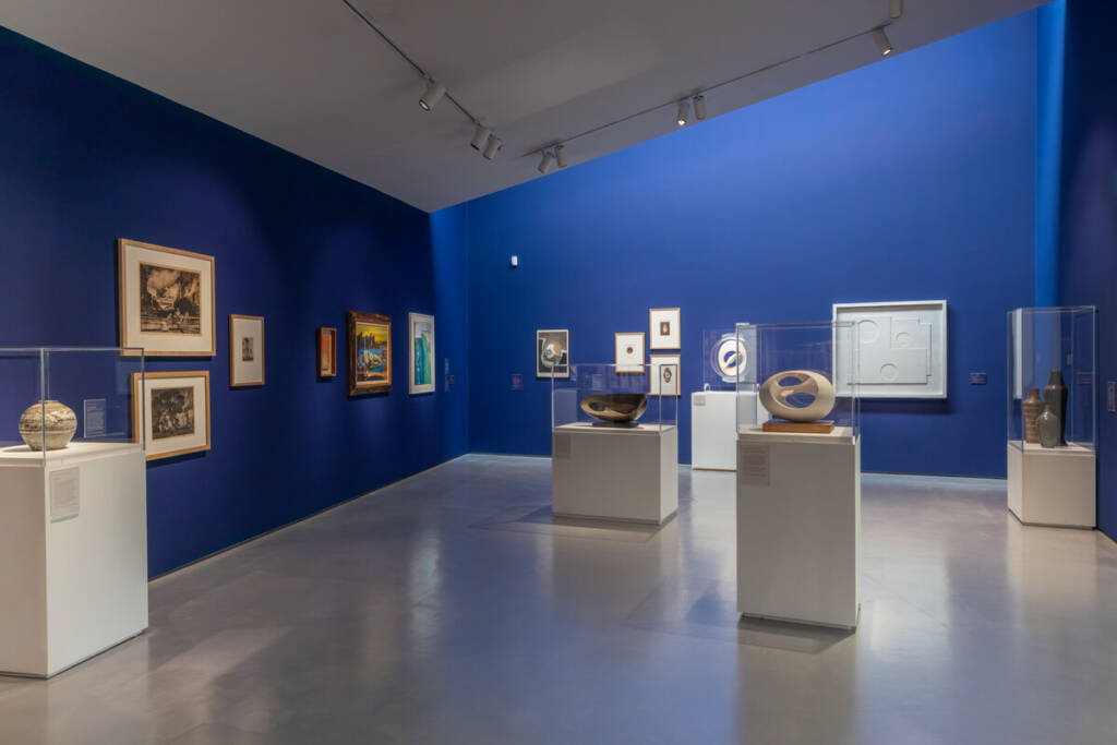 The image shows a modern art gallery with deep blue walls. Various artworks, including paintings and sculptures, are displayed. The room is well-lit, with spotlights highlighting each piece. The floor is a sleek, reflective surface, adding to the contemporary feel of the space.